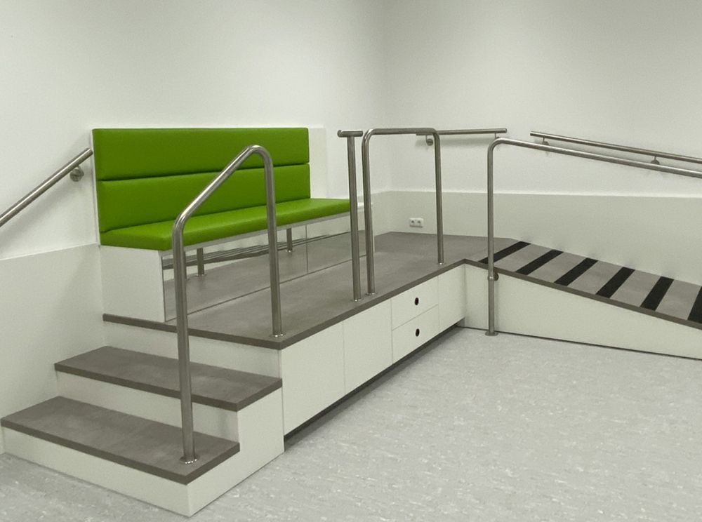 Maßgefertigtes Podest für den barrierefreien Zugang zur Anprobe orthopädischer Hilfsmittel. Das Podest ist weiß, mit einer Lauffläche in Betonoptik und einem stabilen Geländer zur sicheren Nutzung. Die Polsterflächen sind in einem freundlichen Grünton gehalten und farblich auf das Gesamtkonzept des Sanitätshauses abgestimmt.